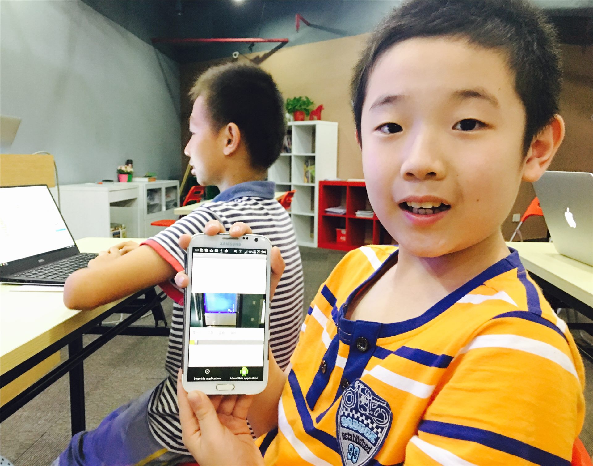 编程课堂中一名学生向镜头展示手机上的应用作品，另一名学生在电脑前学习